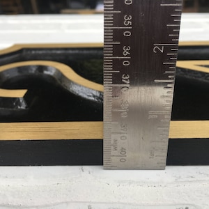 May include: Close-up of a black and gold house number sign with a gold border. A ruler is placed next to the sign to show the size. The ruler measures 37.0 cm.