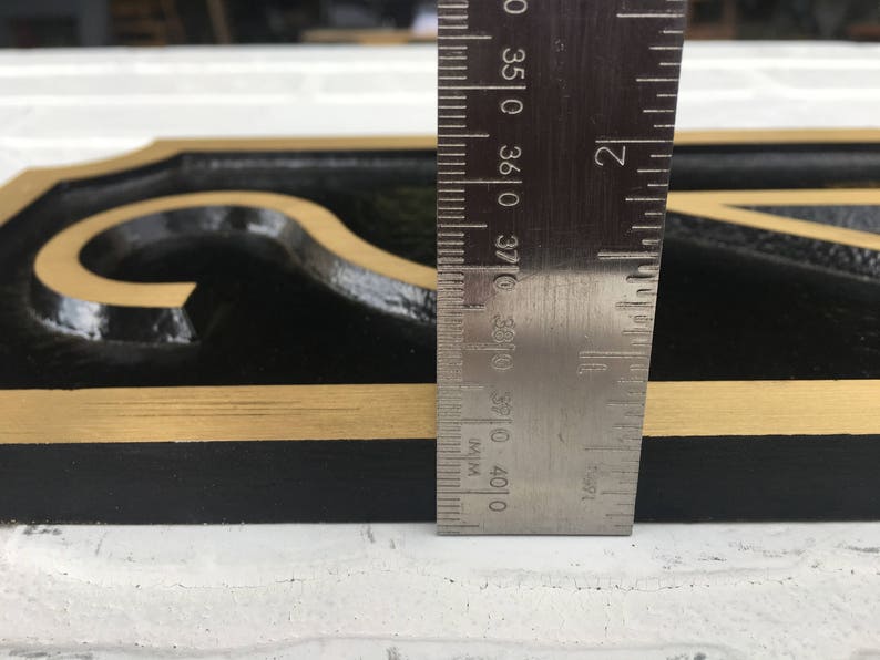May include: Close-up of a black and gold house number sign with a gold border. A ruler is placed next to the sign to show the size. The ruler measures 37.0 cm.