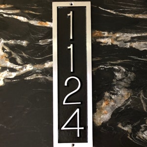 May include: A vertical silver metal house number plaque with a black background. The numbers 1124 are displayed in white.