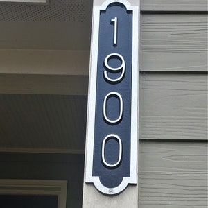 May include: A black and white house number sign with the number 1900 on it. The sign is mounted on a white post.