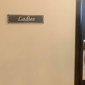 May include: A black door with a brass door handle and a sign that reads "Ladies" above it.