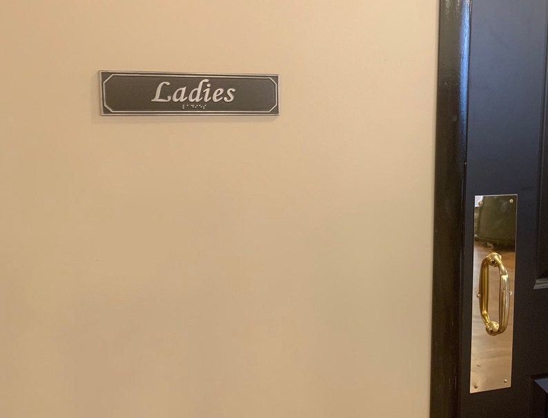 May include: A black door with a brass door handle and a sign that reads "Ladies" above it.
