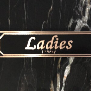 May include: A black rectangular sign with a gold border and the word "Ladies" written in gold cursive lettering.