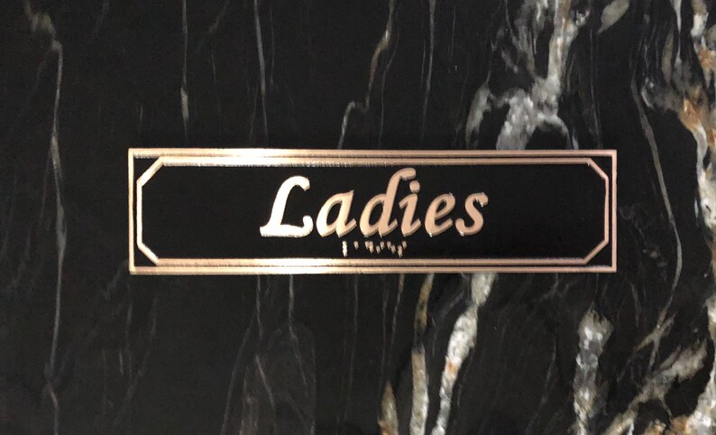 May include: A black rectangular sign with a gold border and the word "Ladies" written in gold cursive lettering.