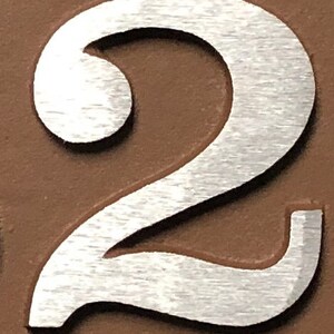 May include: A silver metal number two with a brushed finish. The number is on a brown background.