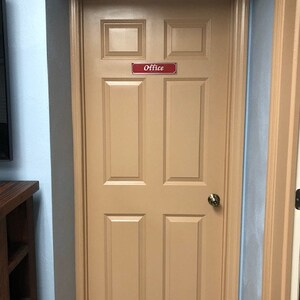 May include: A tan wooden door with a red sign that says "Office" on it. The door has six panels and a brass door knob.