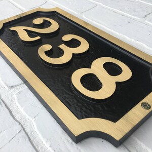 May include: A black and gold house number sign with the numbers 238 in gold lettering. The sign has a decorative border and is mounted on a white brick wall.