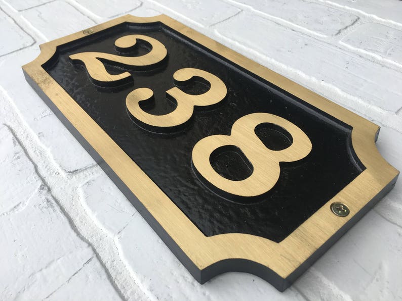 May include: A black and gold house number sign with the numbers 238 in gold lettering. The sign has a decorative border and is mounted on a white brick wall.