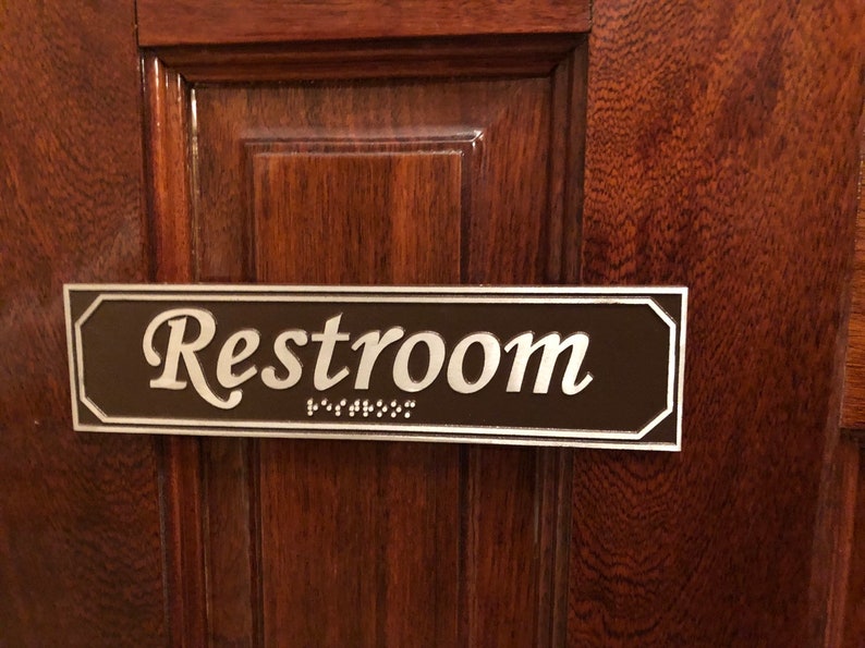 May include: A brown rectangular sign with silver lettering that reads "Restroom" with braille underneath.
