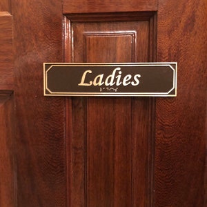 May include: A brown wooden door with a black and gold sign that reads "Ladies" in gold lettering. The sign has braille lettering below the word "Ladies".