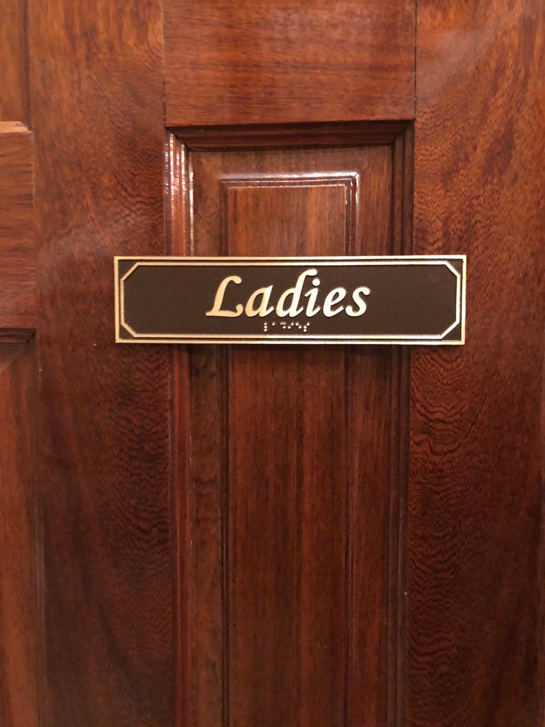 May include: A brown wooden door with a black and gold sign that reads "Ladies" in gold lettering. The sign has braille lettering below the word "Ladies".