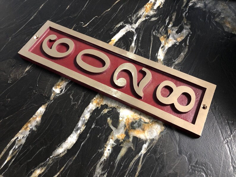 May include: A rectangular brass house number plaque with a red background. The numbers "8028" are cut out of the brass and are raised against the red background.