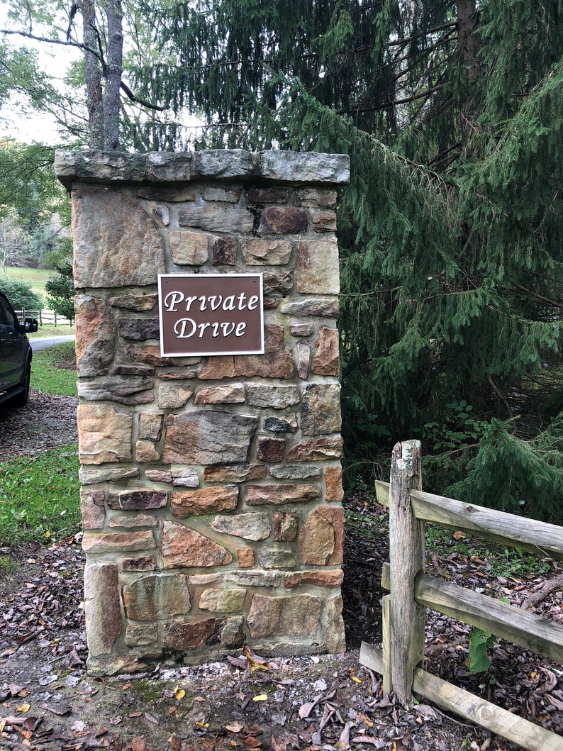 以下が含まれることがあります： 「Private Drive」と書かれた茶色の看板がついた石の柱。柱は積み重ねられた石でできており、緑の木々と木製のフェンスに囲まれている。