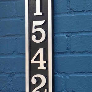 K&ouml;nnte beinhalten: Ein vertikales, ovales Hausnummernschild mit schwarzem Hintergrund und silbernen Zahlen. Die Zahlen sind 1542.