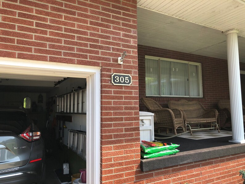 May include: A brick house with a garage door and a porch. The house number 305 is displayed on the front of the house. There are two wicker chairs and a wicker sofa on the porch.