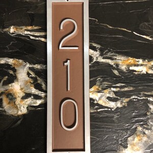 May include: A brown rectangular house number sign with a silver frame. The number 210 is displayed in white on the sign.