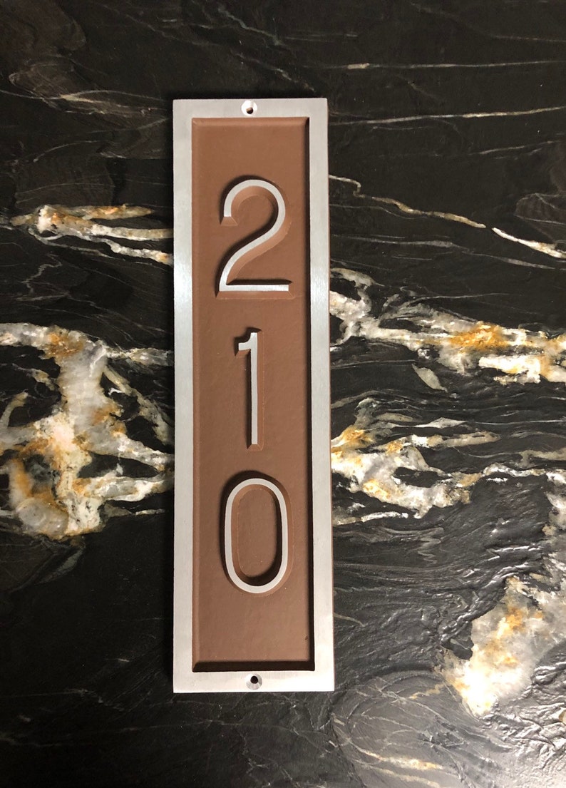 May include: A brown rectangular house number sign with a silver frame. The number 210 is displayed in white on the sign.