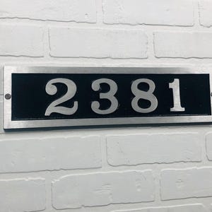 May include: A rectangular address plaque with the number "2381" in silver against a black background. The plaque is framed in silver and mounted on a white brick wall. The numbers are large and easy to read.