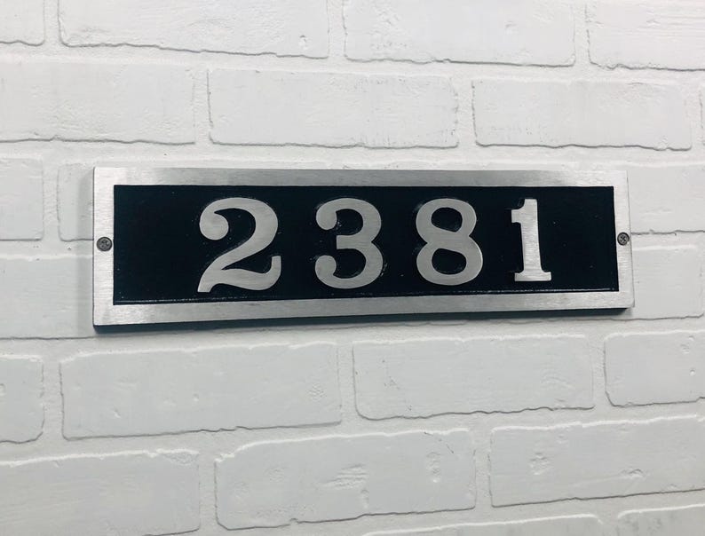 May include: A rectangular address plaque with the number "2381" in silver against a black background. The plaque is framed in silver and mounted on a white brick wall. The numbers are large and easy to read.