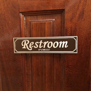 May include: A brown wooden door with a silver plaque that reads "Restroom" in cursive lettering. The plaque also has braille lettering below the word "Restroom".