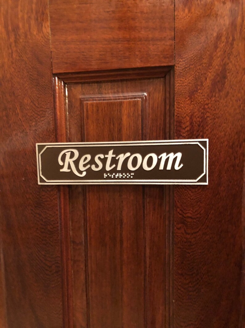 May include: A brown wooden door with a silver plaque that reads "Restroom" in cursive lettering. The plaque also has braille lettering below the word "Restroom".