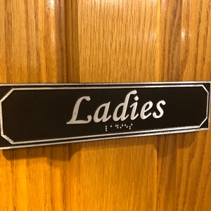 May include: A black rectangular sign with silver trim and the word "Ladies" in silver lettering. The sign also includes braille lettering.