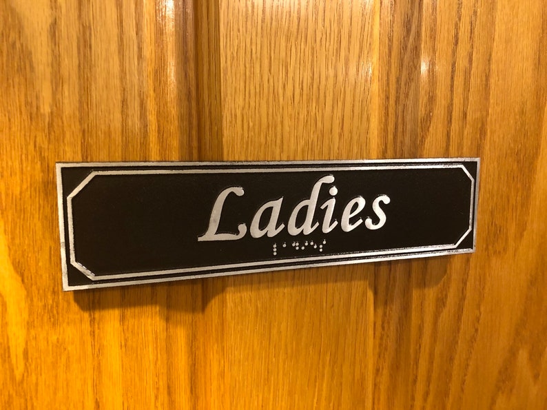 May include: A black rectangular sign with silver trim and the word "Ladies" in silver lettering. The sign also includes braille lettering.