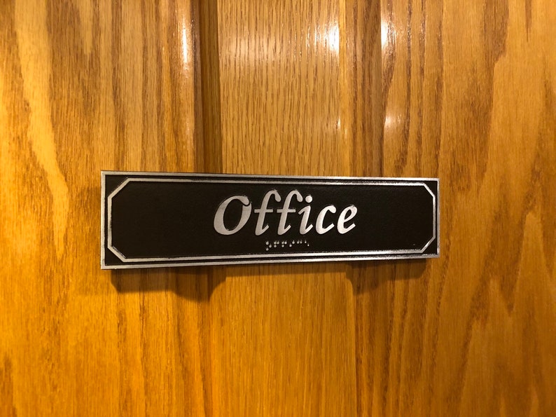 May include: A black and silver rectangular sign with the word "Office" in silver lettering. The sign also includes braille lettering.