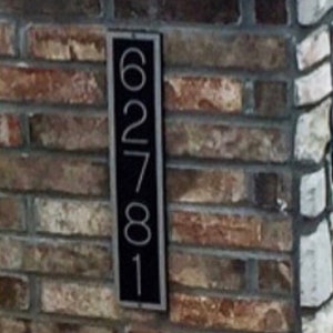 May include: A black metal house number plaque with the numbers 62781 on it. The plaque is mounted on a brick wall.