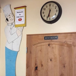 Pu&ograve; includere: Un ritaglio di chef dei cartoni animati con un cartello che dice "Benvenuti da Chili Joe's". C'&egrave; un orologio sul muro dietro lo chef e una porta di legno con un cartello che dice "Bagno".