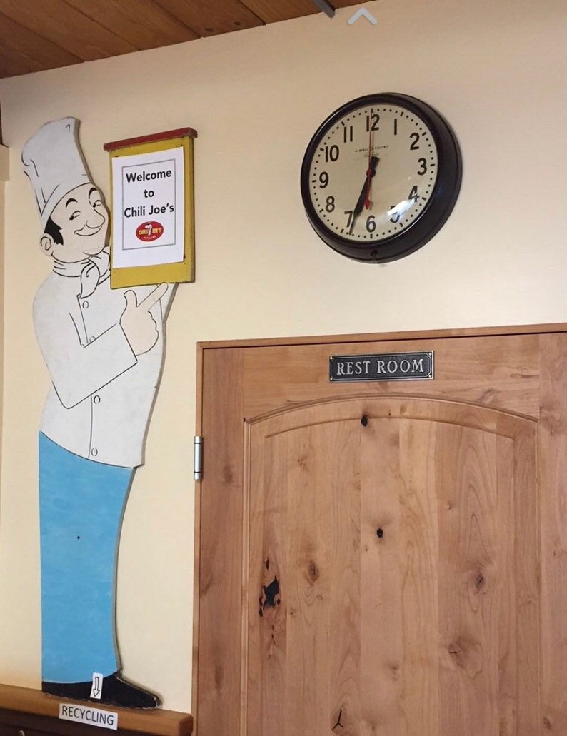 Pu&ograve; includere: Un ritaglio di chef dei cartoni animati con un cartello che dice "Benvenuti da Chili Joe's". C'&egrave; un orologio sul muro dietro lo chef e una porta di legno con un cartello che dice "Bagno".
