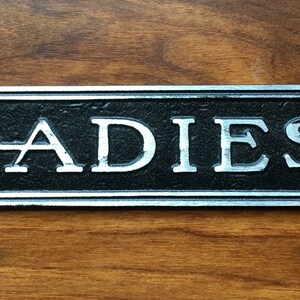 K&ouml;nnte beinhalten: Ein silbernes Metallschild mit schwarzer Beschriftung, auf dem "LADIES" steht.