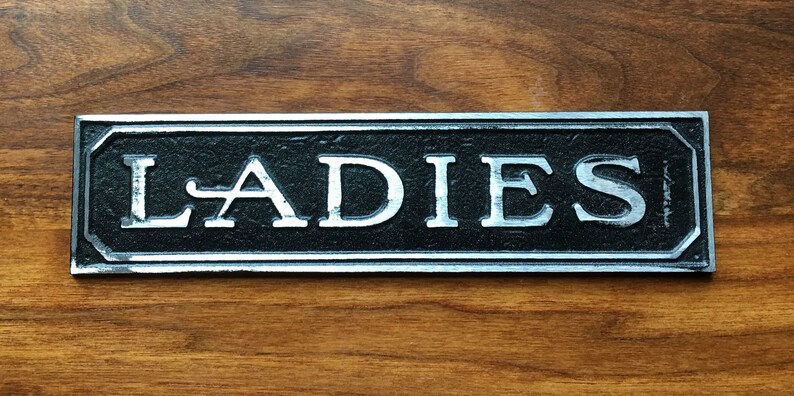 K&ouml;nnte beinhalten: Ein silbernes Metallschild mit schwarzer Beschriftung, auf dem "LADIES" steht.