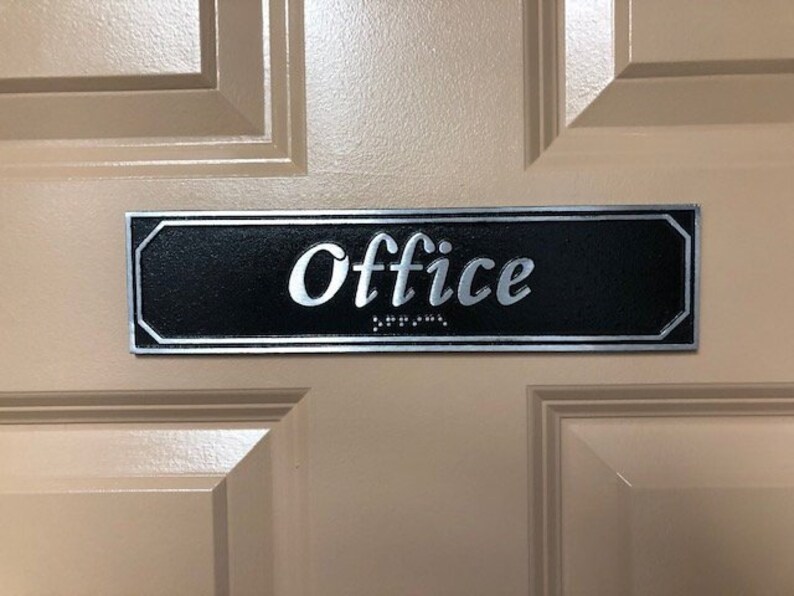 May include: A black and silver rectangular sign with the word "Office" in silver lettering. The sign has braille lettering below the word "Office".