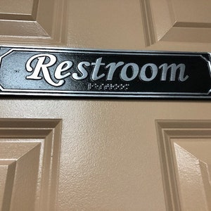 May include: A black and silver sign with the word "Restroom" in a decorative script font.
