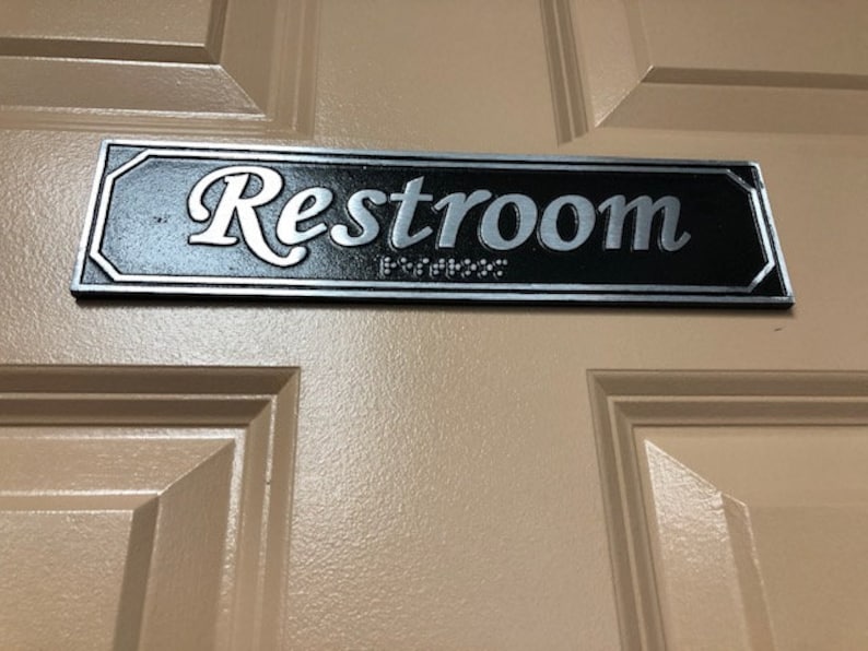 May include: A black and silver sign with the word "Restroom" in a decorative script font.