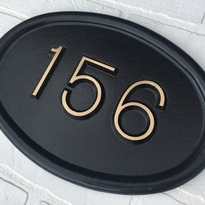 May include: Black oval-shaped house number plaque with gold-colored numbers "156" on a white brick wall background.