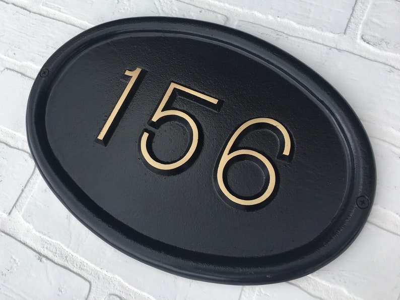May include: Black oval-shaped house number plaque with gold-colored numbers "156" on a white brick wall background.