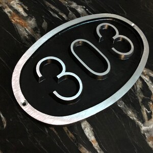 May include: A silver oval-shaped sign with a black background and the number 308 in raised silver lettering.