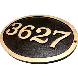 May include: Oval-shaped house number plaque with a black background and gold-coloured numbers. The numbers "3627" are raised and visible against the black background.