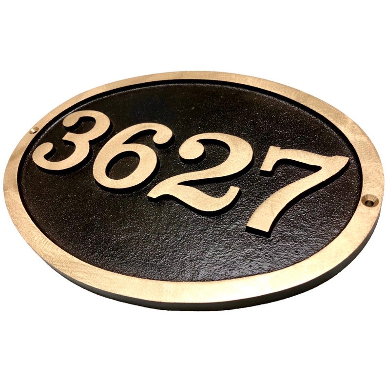 May include: Oval-shaped house number plaque with a black background and gold-coloured numbers. The numbers "3627" are raised and visible against the black background.