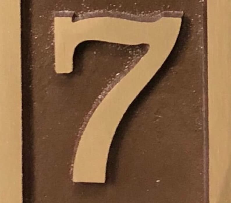 May include: A close-up of a bronze-coloured house number plaque with the number 7 embossed in a lighter shade of gold.
