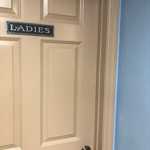 May include: A white door with a black sign that says "LADIES" on it. The door has a silver door knob and is framed by a brown trim. The wall next to the door is light blue.