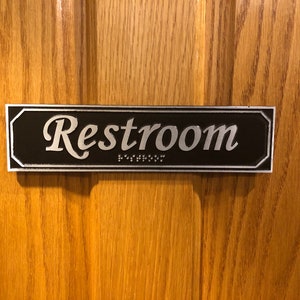 May include: A black and silver rectangular sign with the word "Restroom" in silver lettering and braille dots below the word.