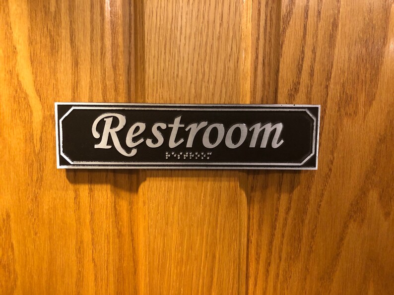 May include: A black and silver rectangular sign with the word "Restroom" in silver lettering and braille dots below the word.