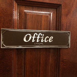 May include: A brown rectangular sign with silver trim and rounded corners. The sign says "Office" in a silver cursive font with braille lettering below.