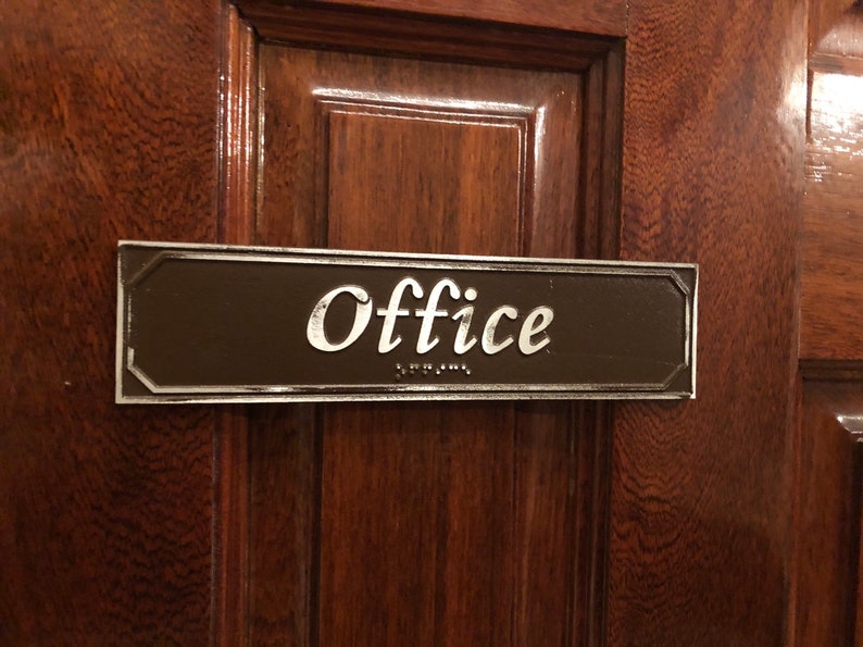 May include: A brown rectangular sign with silver trim and rounded corners. The sign says "Office" in a silver cursive font with braille lettering below.