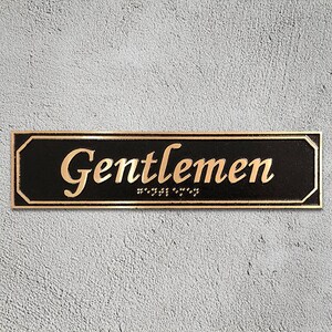 May include: A black rectangular sign with gold lettering that reads "Gentlemen" with braille underneath.