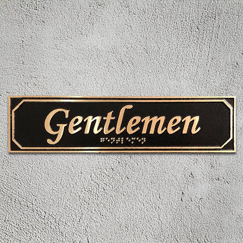 May include: A black rectangular sign with gold lettering that reads "Gentlemen" with braille underneath.