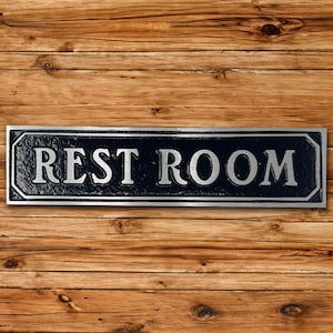 Pu&ograve; includere: Un cartello in metallo nero e argento con la scritta "REST ROOM" in lettere argentate.